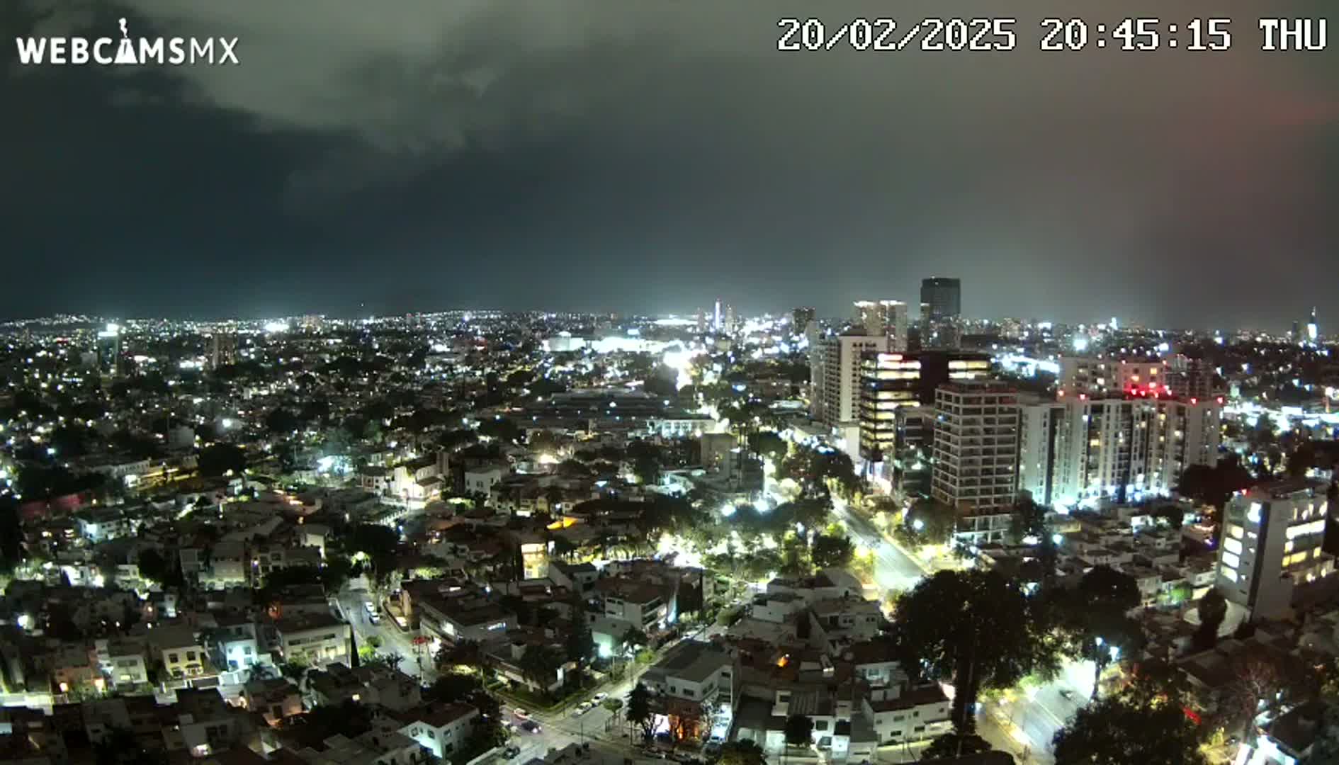 A nighttime, high-angle view of a city under a partly cloudy sky shows many buildings with lights on.