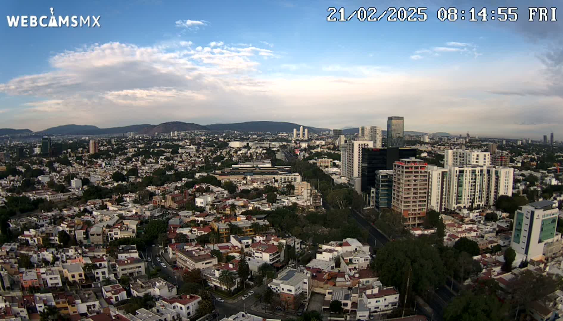 An aerial view of a city on a partly cloudy day, showing a mix of residential buildings and taller structures, with mountains visible in the distance.