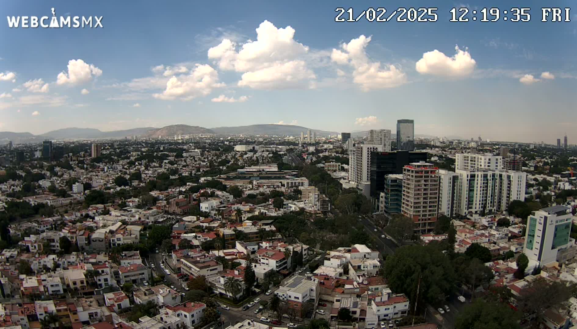 An aerial view of a sprawling city under a partly cloudy sky shows numerous residential buildings and taller structures, with mountains visible in the distance.