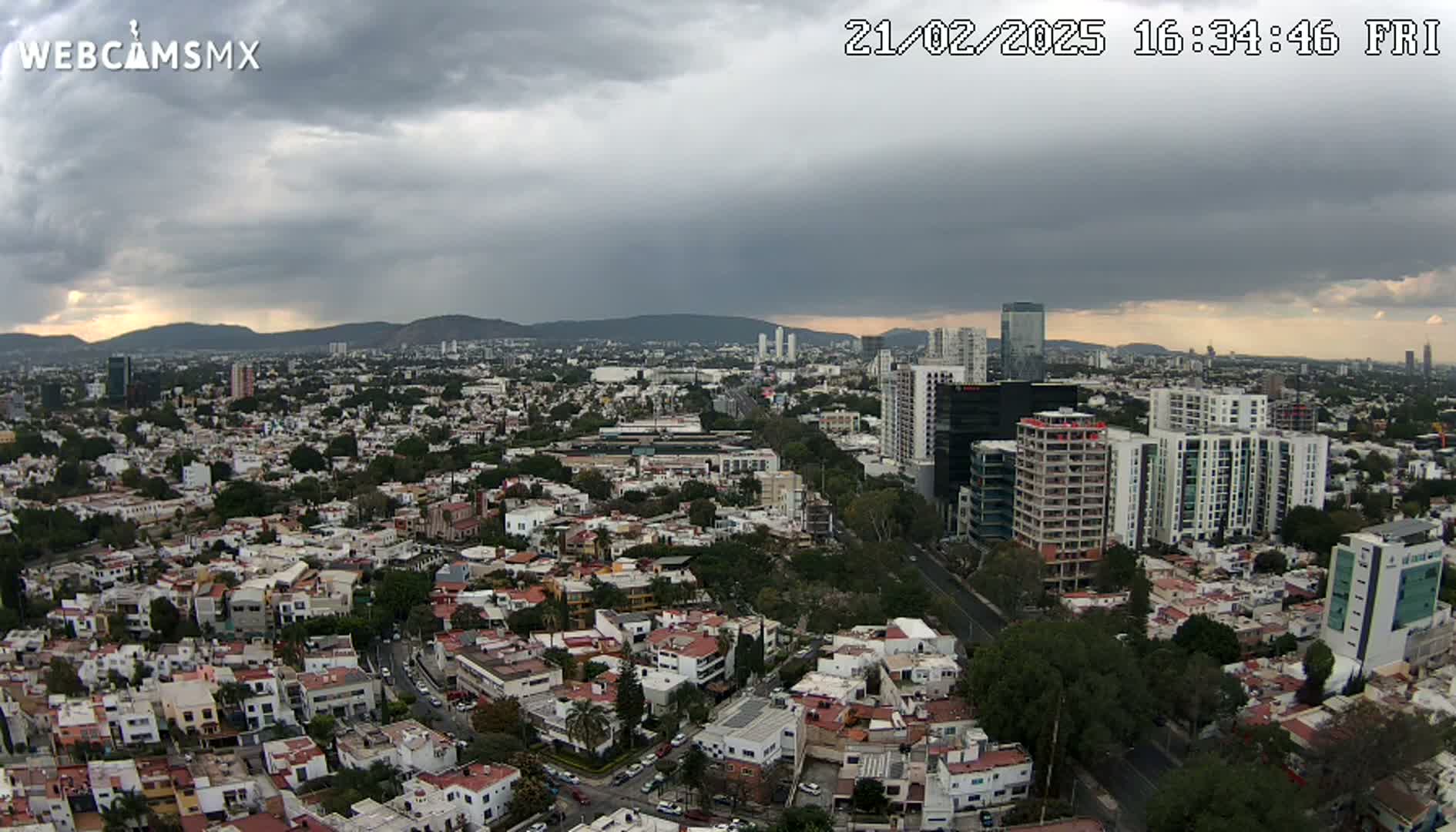 An expansive cityscape under a cloudy sky shows a sprawling network of residential buildings and taller structures, nestled amongst green spaces and roads.