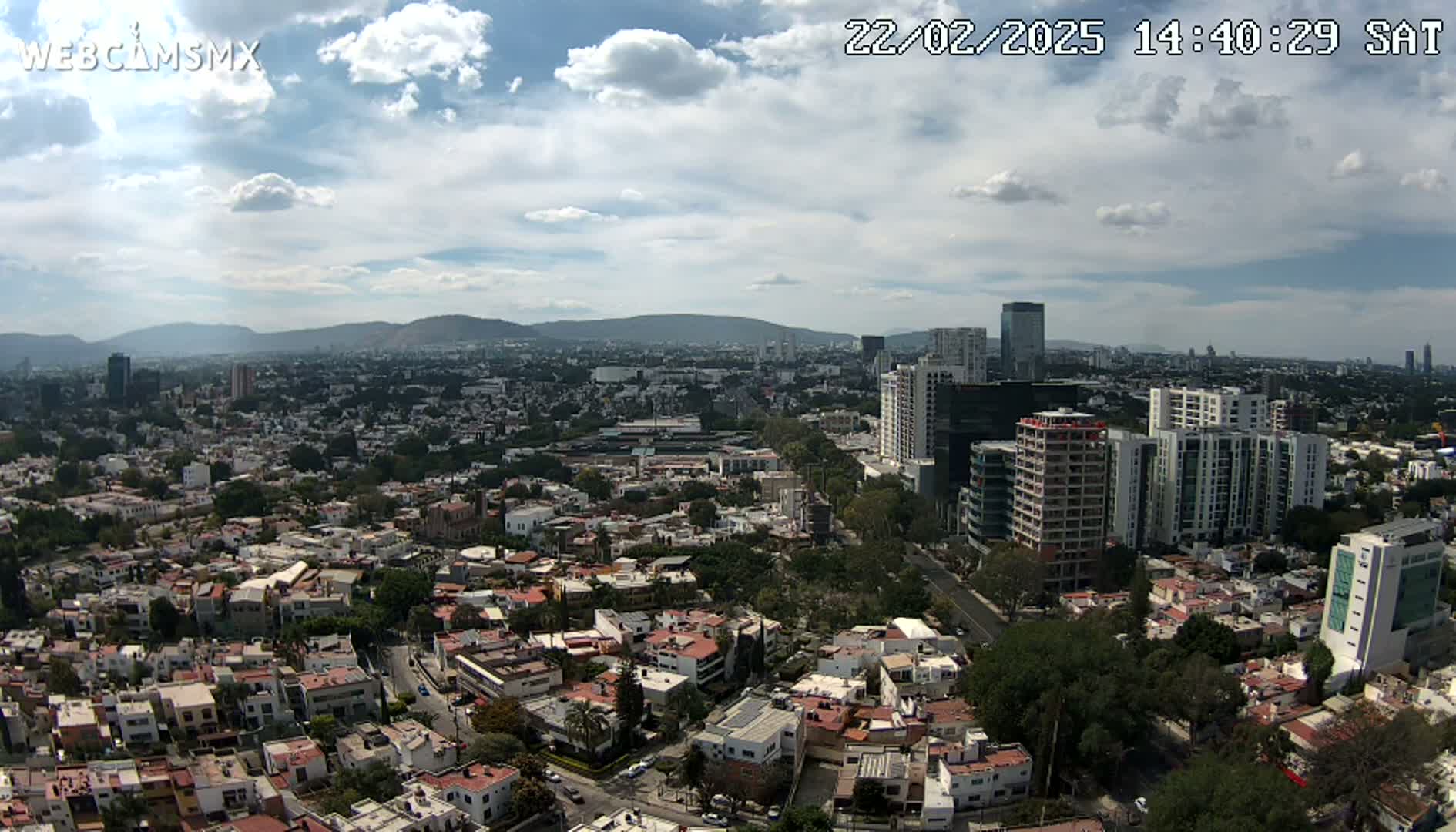 An aerial view shows a sprawling city under a partly cloudy sky, with mountains visible in the distance.