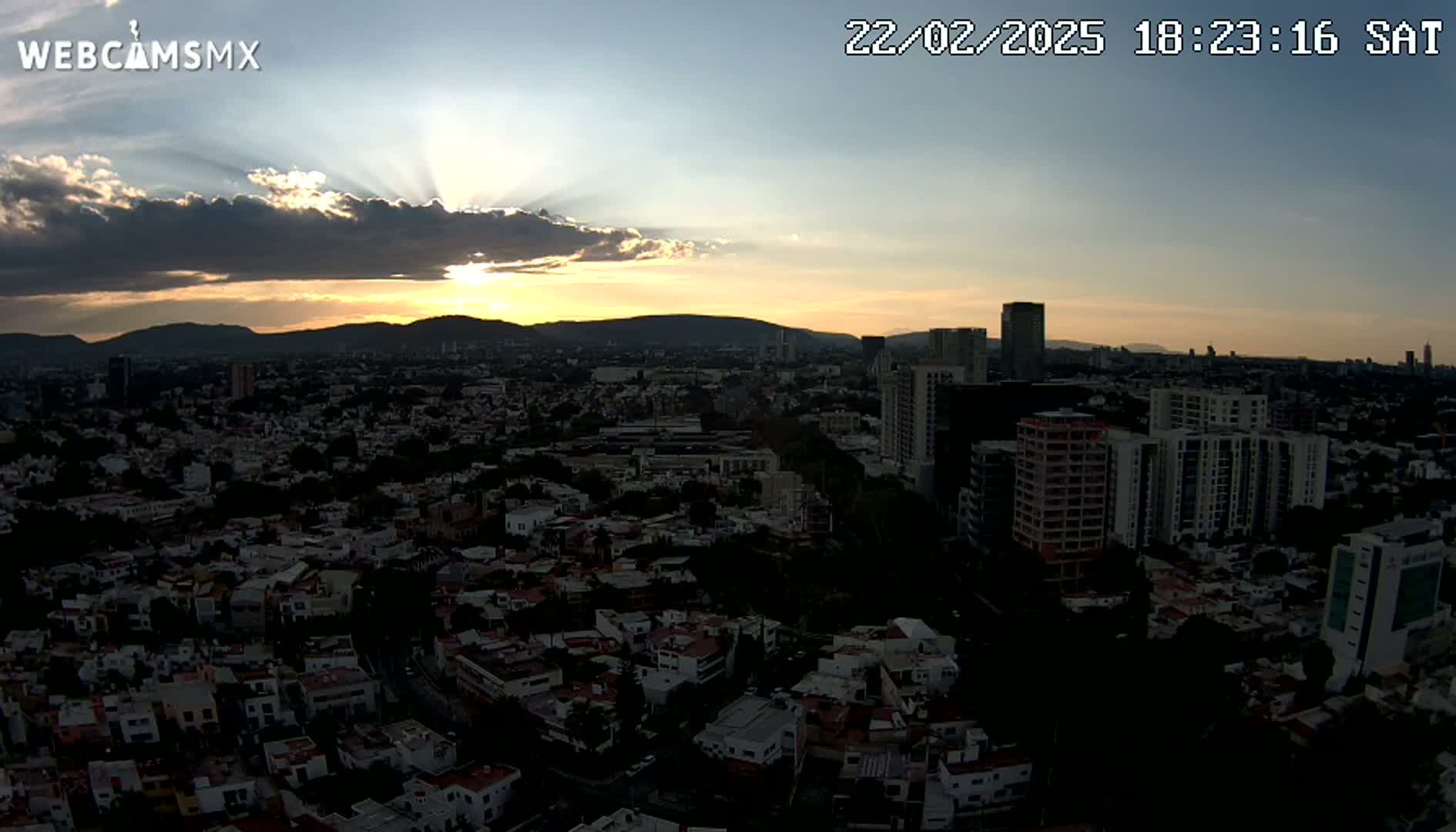 A sprawling city at sunset, with crepuscular rays emanating from behind a cloud layer over distant mountains.
