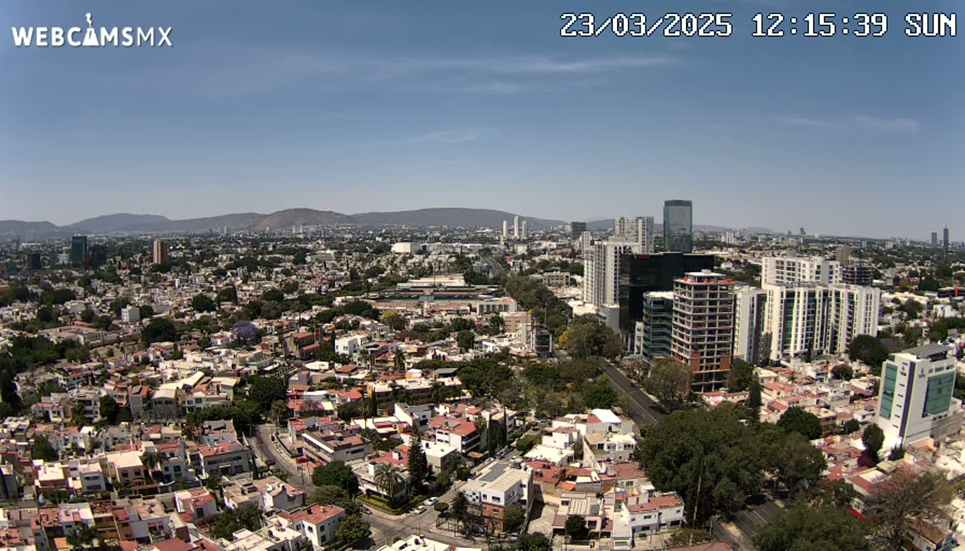 A sunny, aerial view of a sprawling city with numerous residential buildings, taller structures, and mountains in the distance.