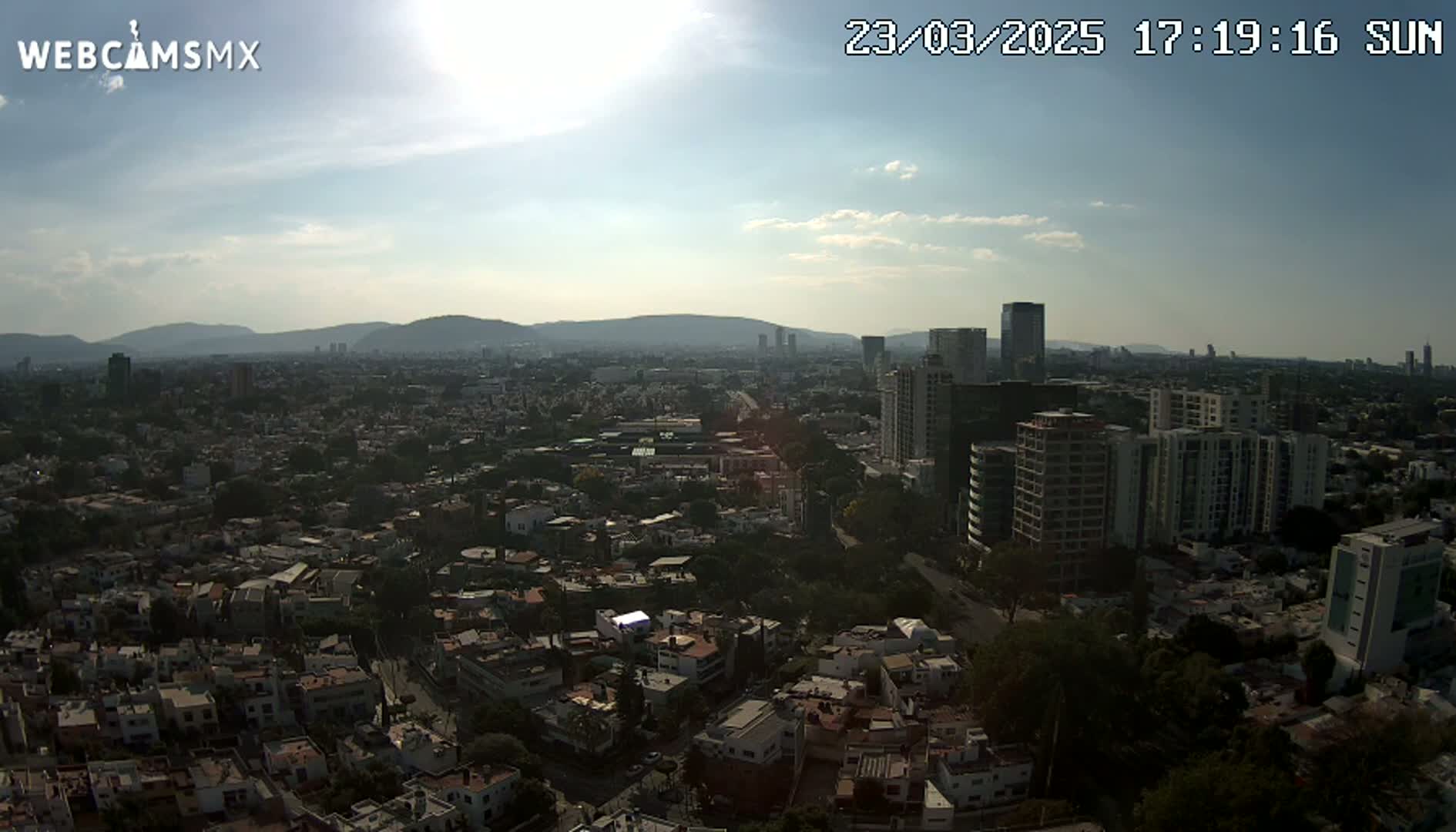 A sunny, partly cloudy aerial view shows a sprawling city with numerous residential buildings and taller structures, backed by distant mountains.