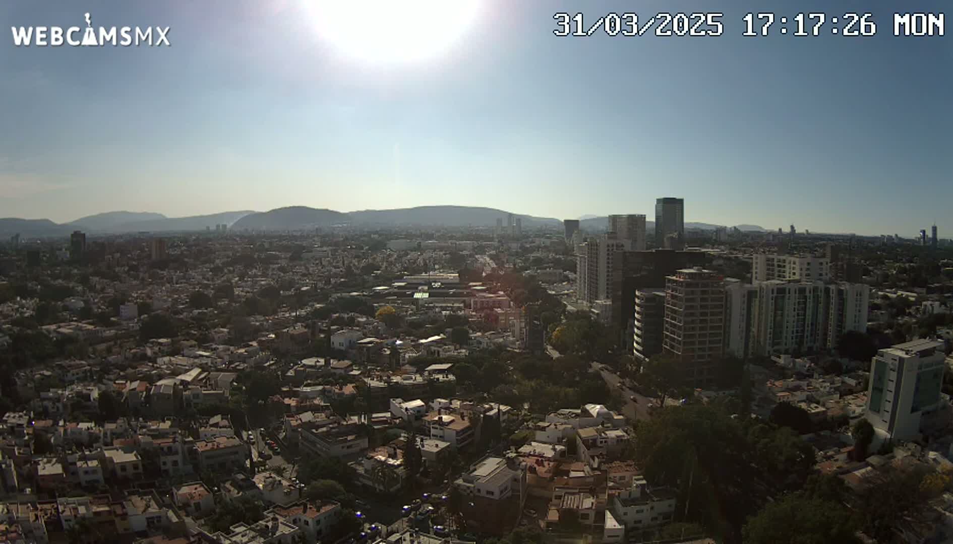 A sunny, aerial view shows a sprawling city with numerous buildings of varying heights, nestled against a backdrop of distant mountains.