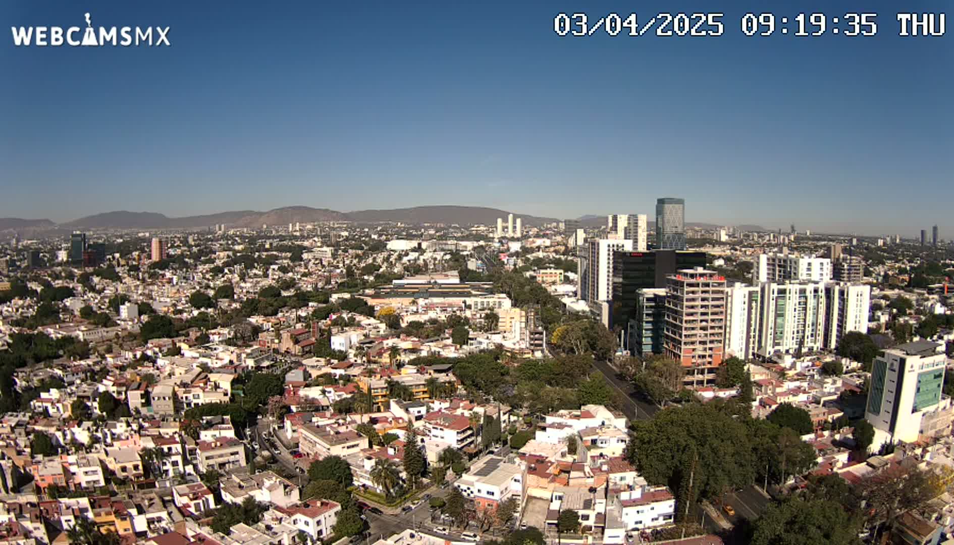An aerial view of a sprawling city on a clear, sunny day, showing a mix of residential buildings and taller structures, with mountains visible in the distance.