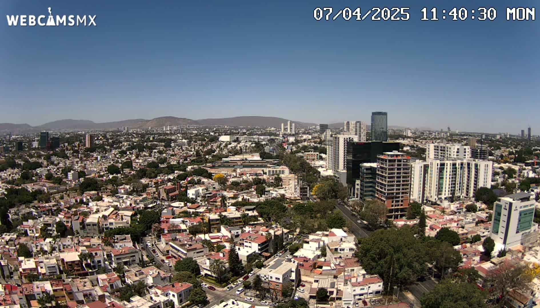 A sprawling cityscape under a clear blue sky, featuring a mix of low-rise residential buildings and taller apartment and office structures, stretches to distant mountains.