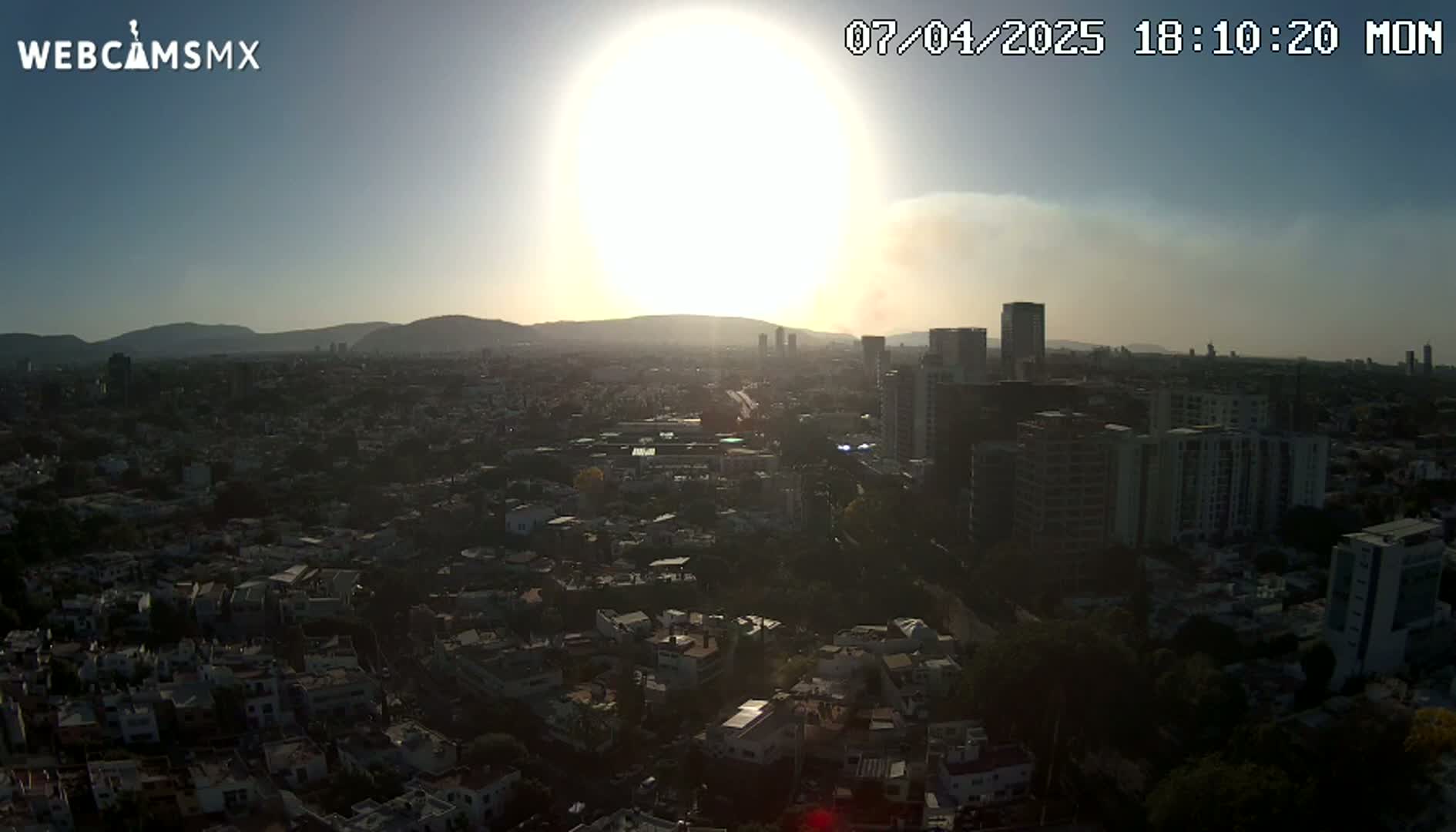 A hazy sunset illuminates a sprawling city, with mountains visible in the distance.
