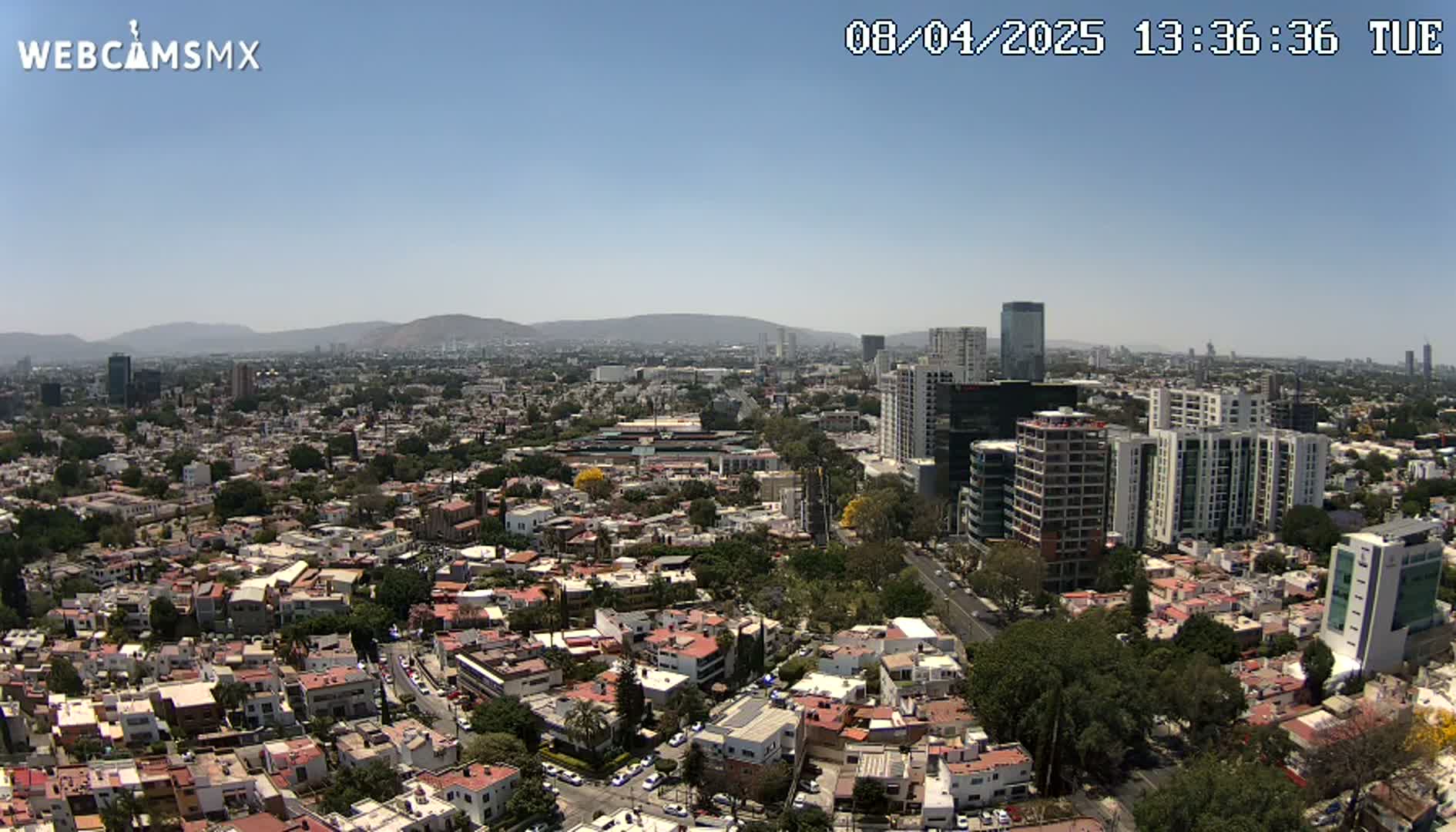 A high-angle, long shot shows a sprawling city under a clear, sunny sky, with mountains visible in the distance.