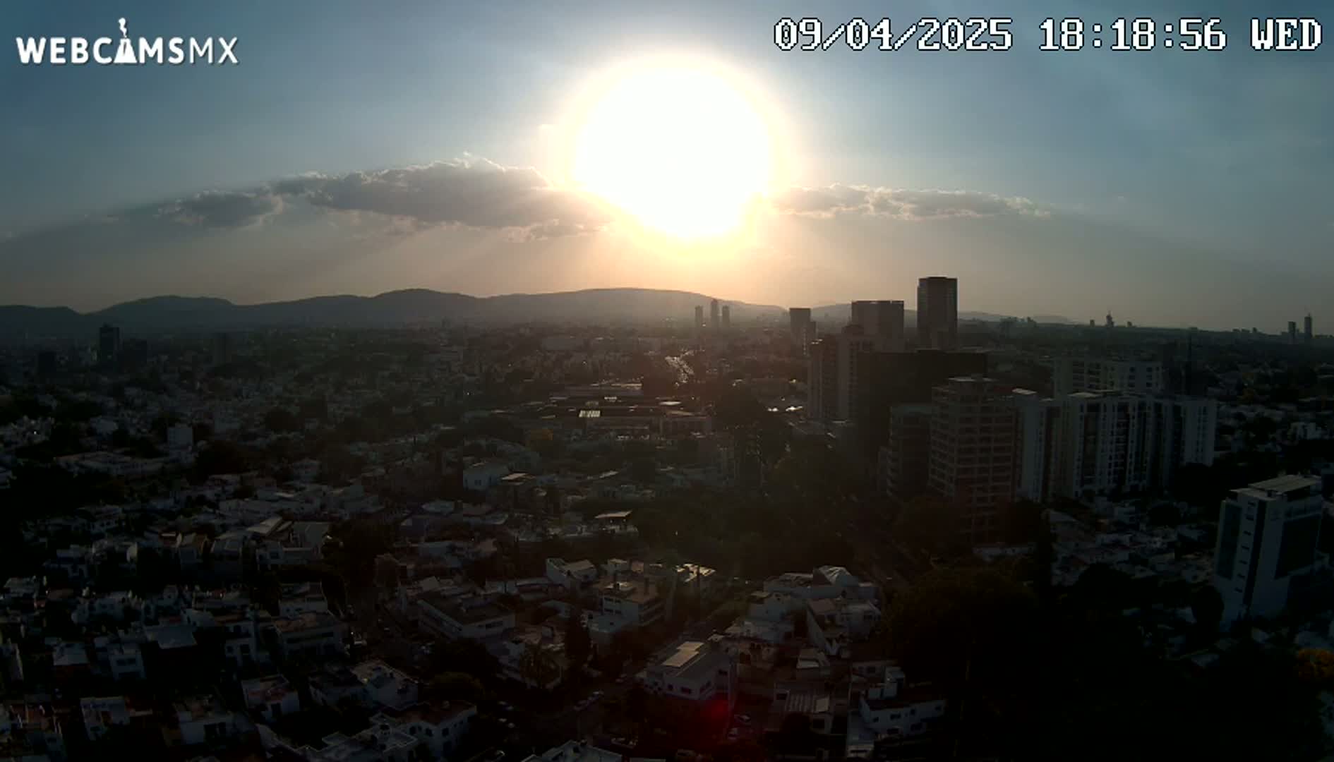 A mostly sunny sunset casts a golden glow over a sprawling city, partially obscured by shadows, with distant mountains visible on the horizon.