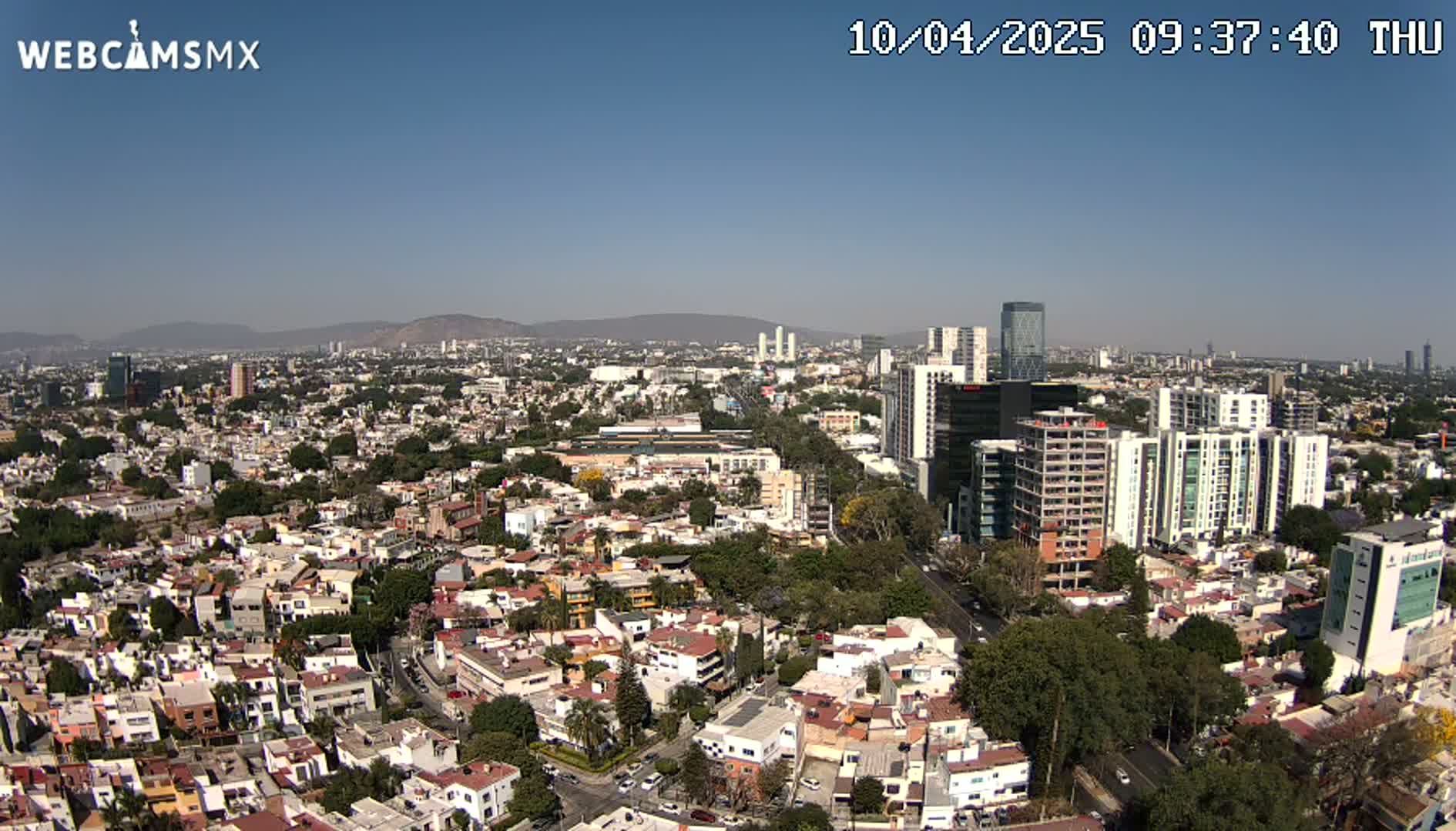 An aerial view of a sprawling city under a clear, sunny sky shows a mix of residential buildings and taller structures, with distant mountains visible on the horizon.