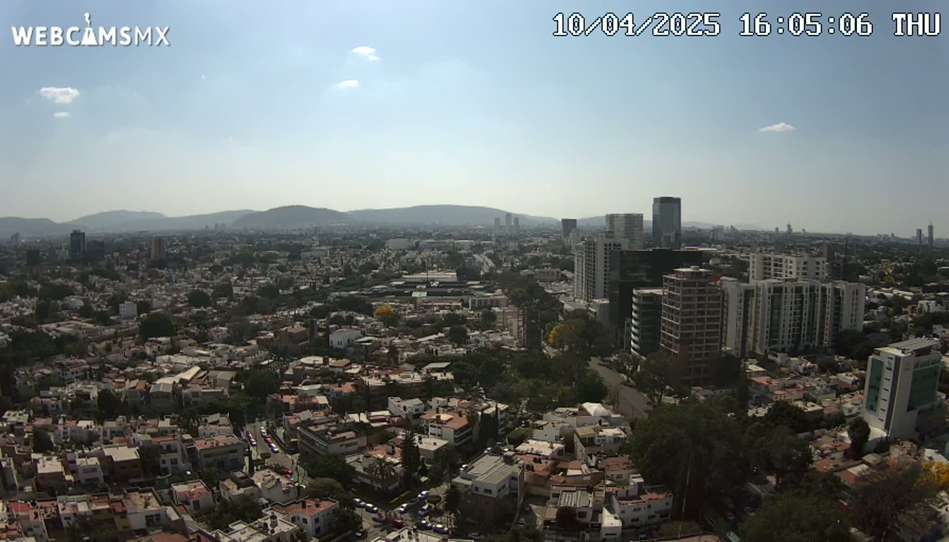 A sunny aerial view shows a sprawling city with numerous residential buildings and taller structures, backed by distant mountains.