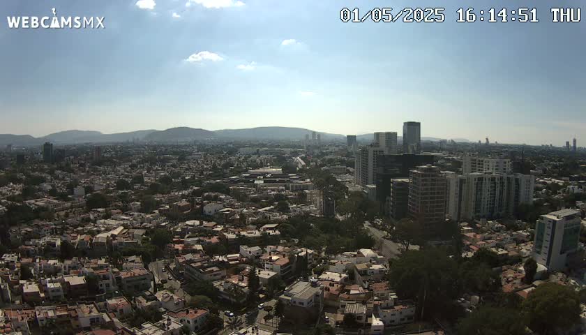 A sprawling city scene under a mostly sunny sky, showing a mix of residential homes and taller buildings, with mountains visible in the distance.