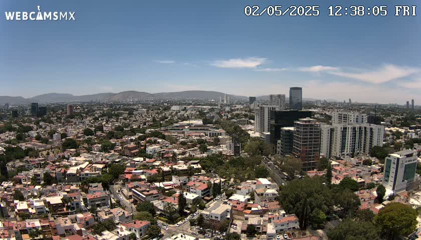 An aerial view on a clear, sunny day shows a sprawling city with low-rise buildings and taller structures interspersed, nestled against a backdrop of distant mountains.