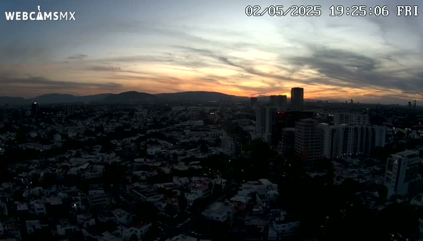 An aerial view of a city at sunset, with a mostly cloudy sky displaying orange and yellow hues.