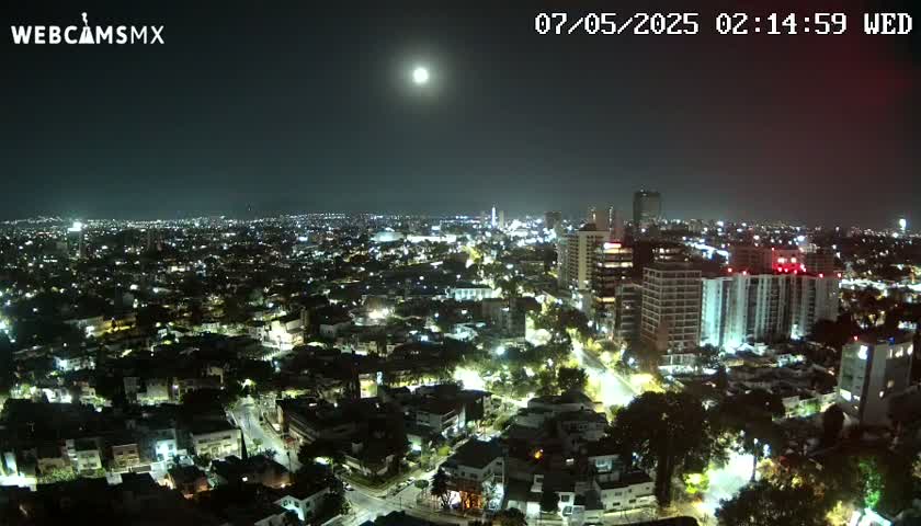 A brightly lit city at night is visible under a clear sky with a full moon.