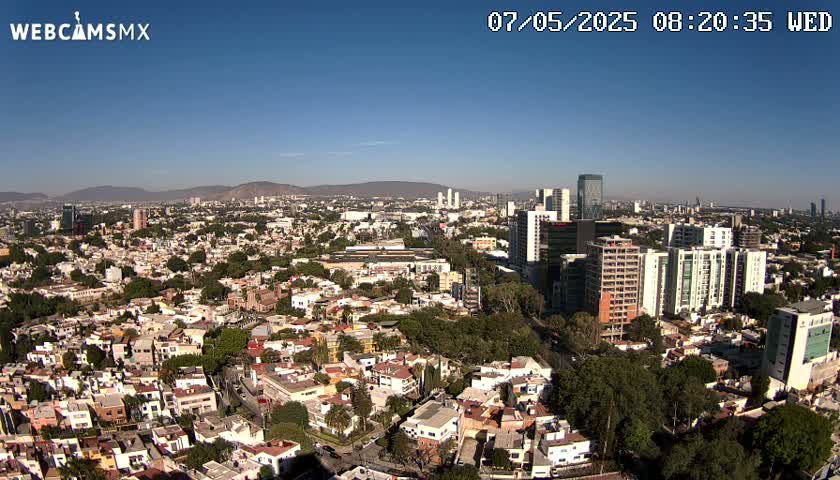 An expansive cityscape with numerous buildings, trees, and mountains in the distance under a clear blue sky.