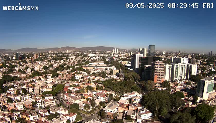 An aerial view of a sprawling city on a clear, sunny day shows a mix of low-rise and high-rise buildings, with mountains visible in the distance.