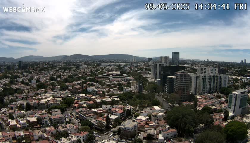 An aerial view of a city on a partly cloudy day shows a sprawling landscape of residential buildings and taller structures, with mountains visible in the distance.