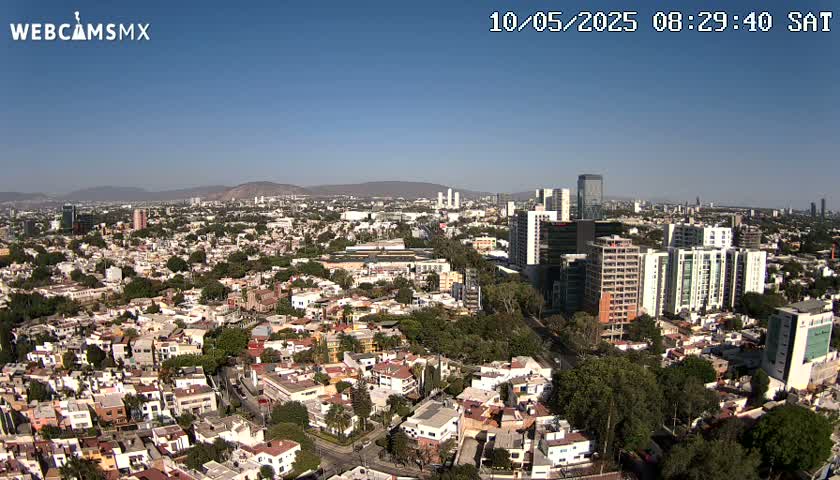A sprawling city landscape under a clear blue sky, showing a mix of low-rise residential buildings and taller structures, extending to distant mountains.