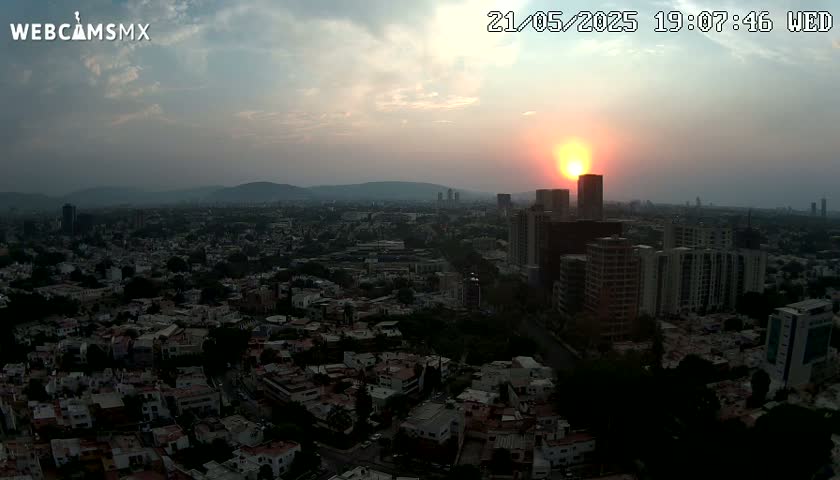A hazy sunset casts an orange glow over a sprawling city of densely packed buildings and low-rise homes, with distant mountains visible on the horizon.