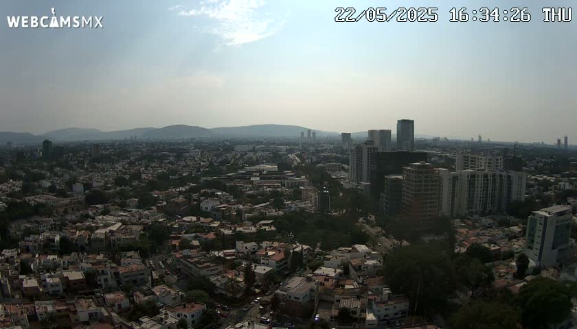 A hazy, sunny day reveals an expansive view of a city's sprawling residential areas and taller buildings, backed by distant mountains.