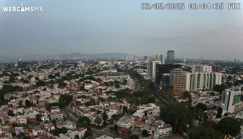 An aerial view of a city at dawn or dusk under hazy skies shows a sprawling landscape of residential neighborhoods and taller buildings in the distance.