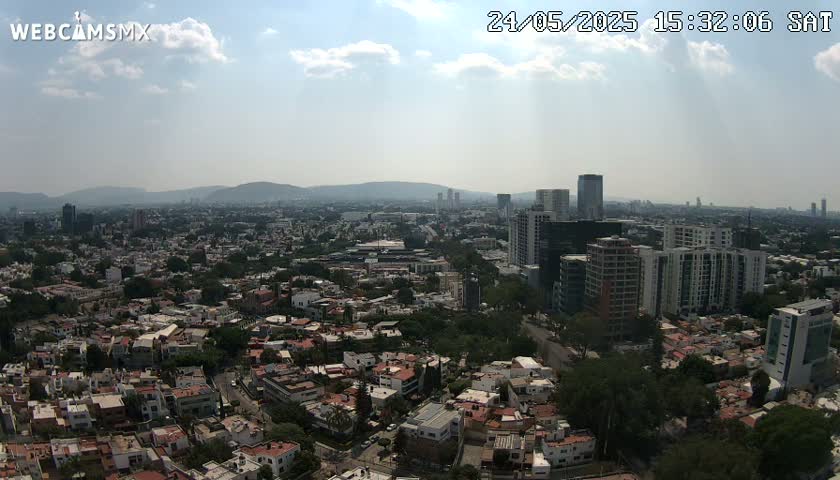 A partly cloudy daytime view of a sprawling city with numerous residential buildings and taller structures, set against a backdrop of distant mountains.