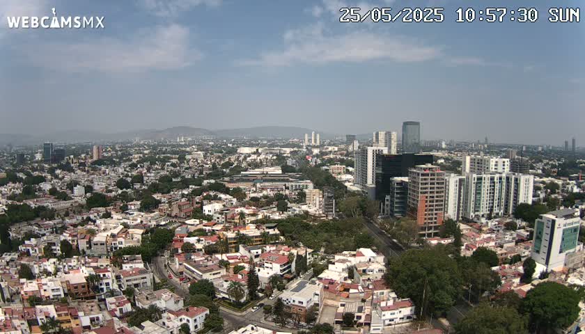 A sprawling city landscape under a mostly clear, sunny sky, showing a mix of low-rise residential buildings and taller modern structures.
