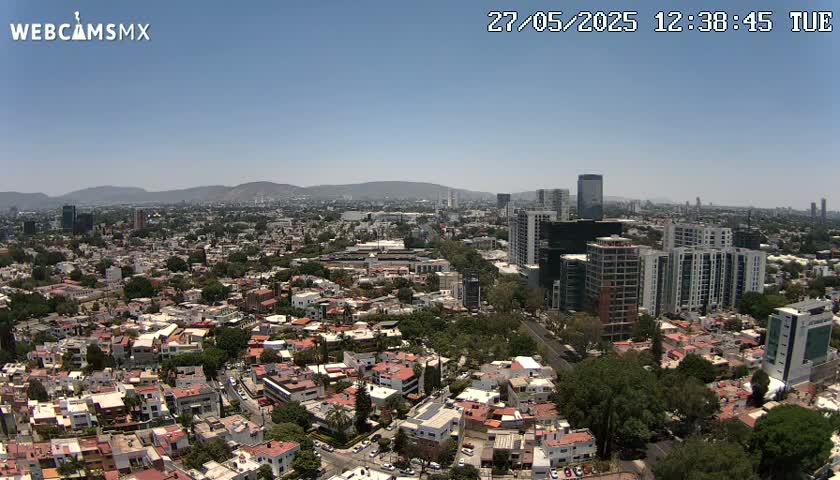 An aerial view shows a sprawling city under a clear blue sky, with mountains visible in the distance.
