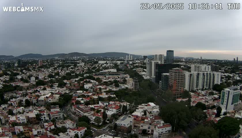 An aerial view of a city under an overcast sky shows numerous residential buildings and taller structures, with mountains visible in the distance.