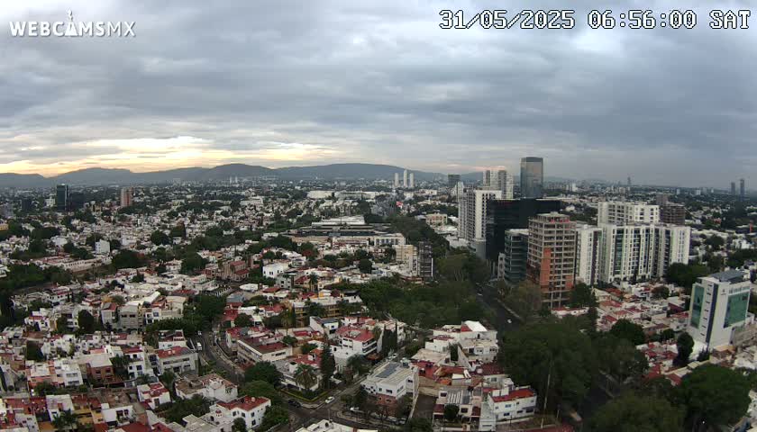 A high-angle, wide shot of a sprawling city under a mostly cloudy sky shows a mix of low-rise and high-rise buildings, with mountains visible in the distance.