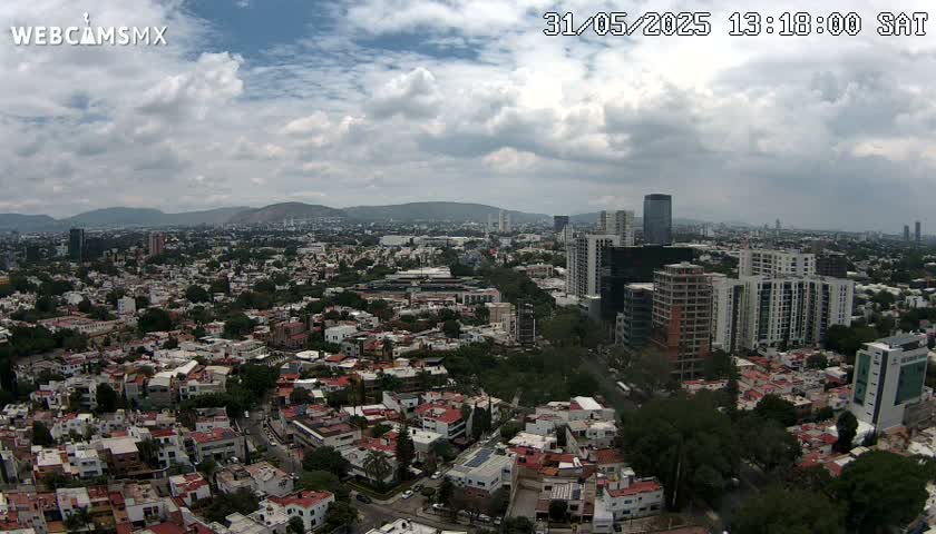 A partly cloudy day reveals a sprawling cityscape of numerous buildings, interspersed with green spaces, extending to distant mountains under a mostly cloudy sky.