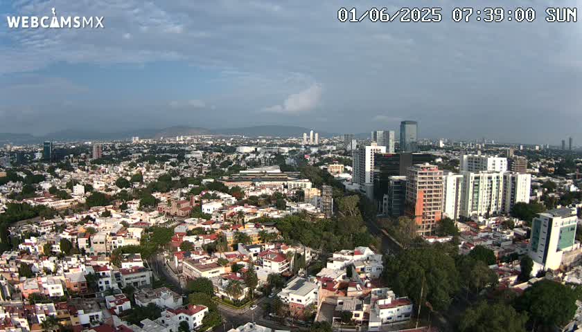 An aerial view of a sprawling city on a partly cloudy day, showing a mix of low-rise residential buildings and taller structures, extending to a skyline in the distance.