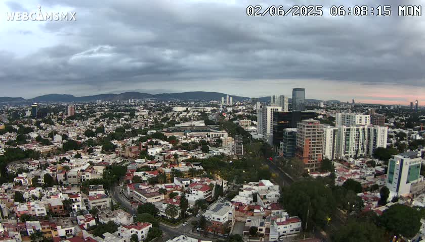 An expansive city view under a cloudy sky shows a mix of low-rise and high-rise buildings extending to distant mountains.