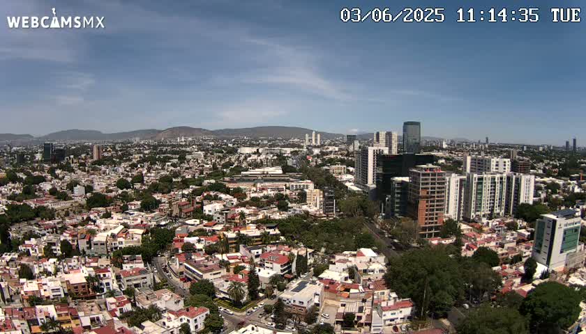 An aerial view of a sprawling city on a partly sunny day, showing a mix of low-rise residential buildings and taller apartment and office structures, with mountains visible in the distance.