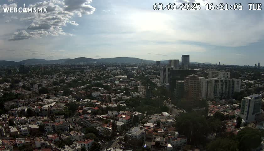 A partly cloudy day reveals a sprawling city with numerous buildings and a mountain range in the distance.
