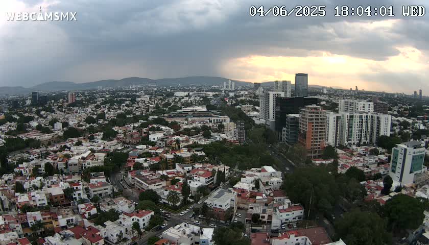 An aerial view of a city under a partly cloudy sky, with the sun setting in the background.