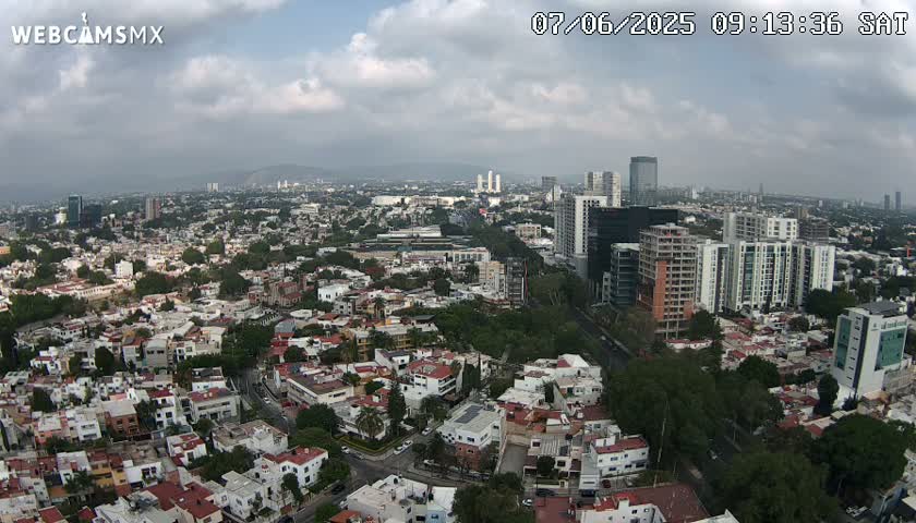 An aerial view of a sprawling city under a partly cloudy sky shows numerous residential buildings and taller structures, including high-rises.
