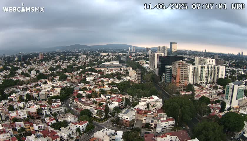An aerial view of a city under a cloudy sky shows a dense collection of residential buildings and taller structures, with a roadway winding through the scene.