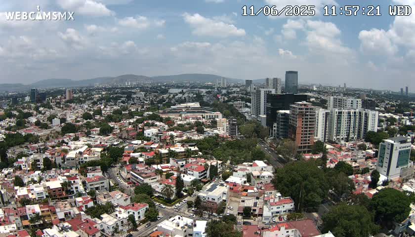 A sprawling cityscape under a partly cloudy sky shows a mix of low-rise residential buildings and taller commercial structures, extending to distant mountains.