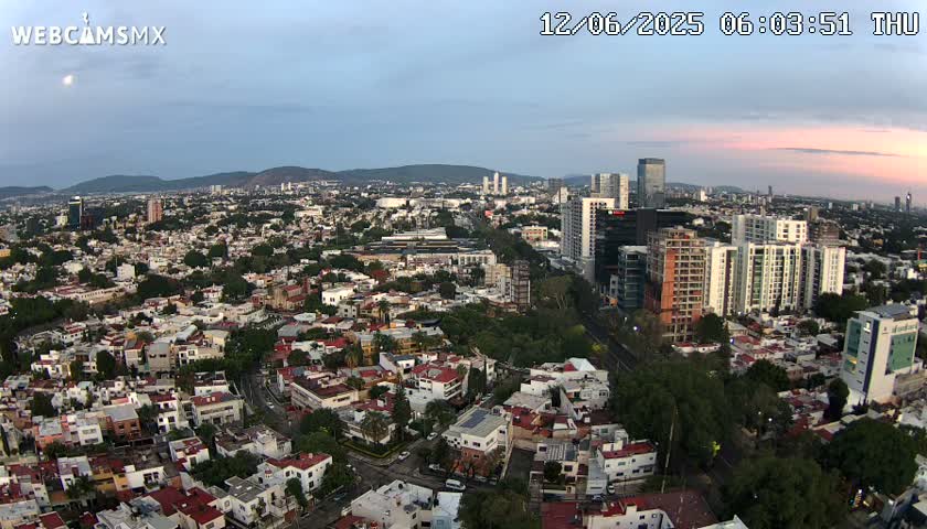 An aerial view of a city at dawn under a mostly clear sky, showing a mix of low-rise residential buildings and taller apartment buildings and skyscrapers.
