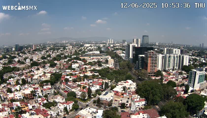 A high-angle, long shot of a city on a partly sunny day shows a dense collection of low-rise residential buildings surrounding taller apartment and office buildings, with mountains visible in the distance.