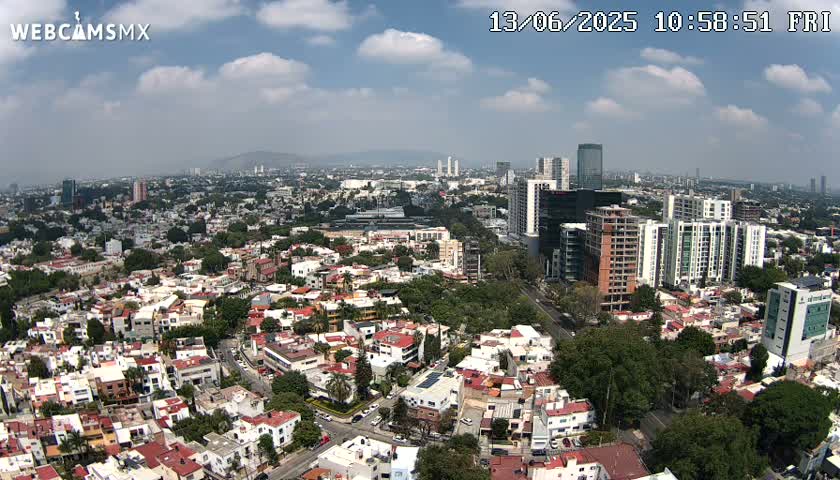 An aerial view of a city on a partly cloudy day shows numerous buildings, including high-rises and low-rise residential structures, interspersed with green spaces.