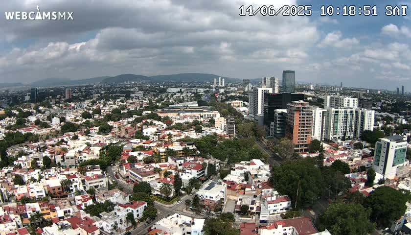 An aerial view of a city on a partly cloudy day shows many residential buildings and taller structures, including several high-rises.