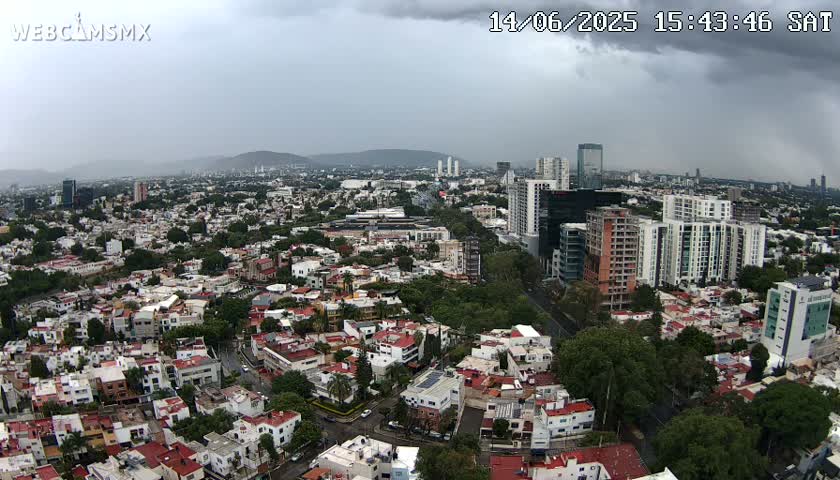 An aerial view of a city on a partly cloudy day shows many residential buildings and taller structures, including several high-rises.