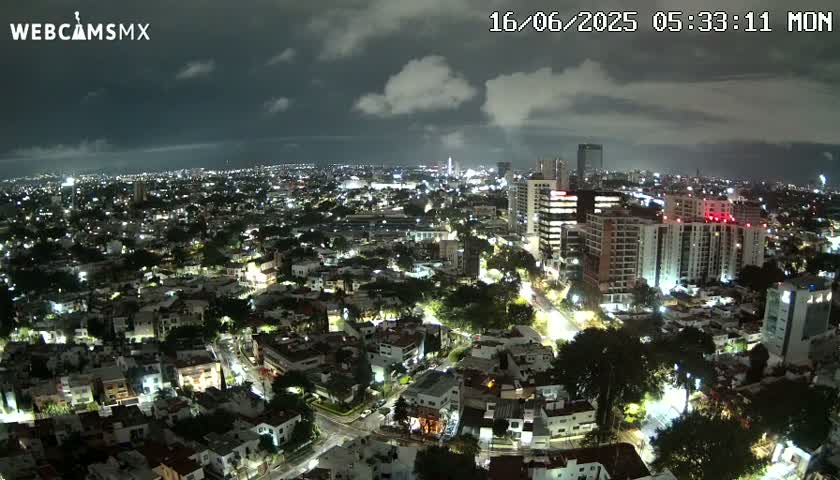 A nighttime aerial view of a city with cloudy skies shows a sprawling cityscape illuminated by streetlights and building lights.