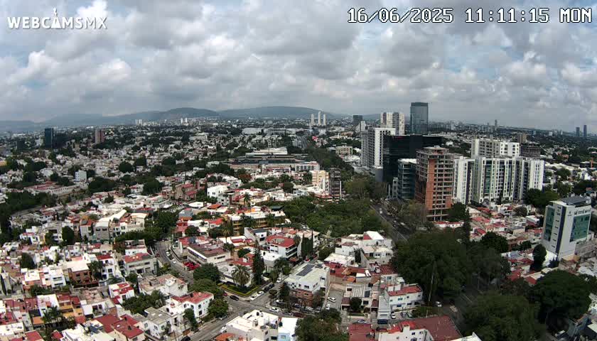 An aerial view of a city on a partly cloudy day, showing a mix of residential buildings and taller structures, with surrounding green areas.