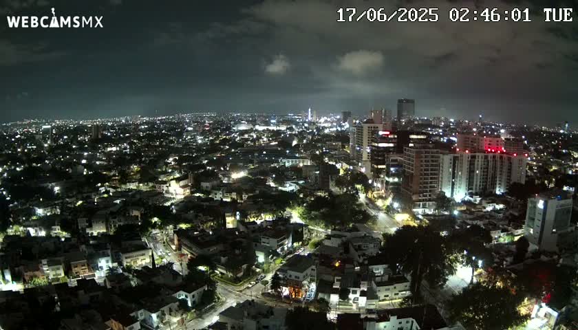 A nighttime aerial view of a city with partly cloudy skies shows numerous illuminated buildings and streets.