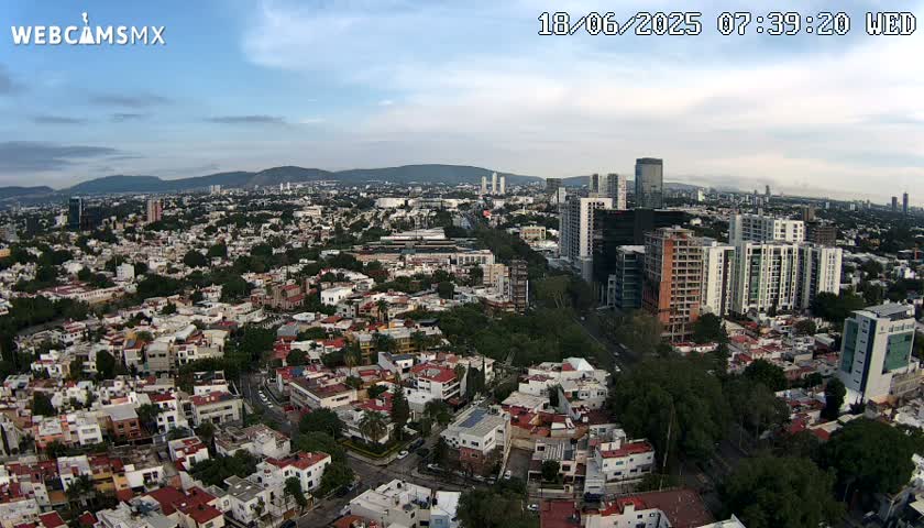 An aerial view of a city under partly cloudy skies, showing a mix of residential buildings and taller structures, with mountains visible in the distance.